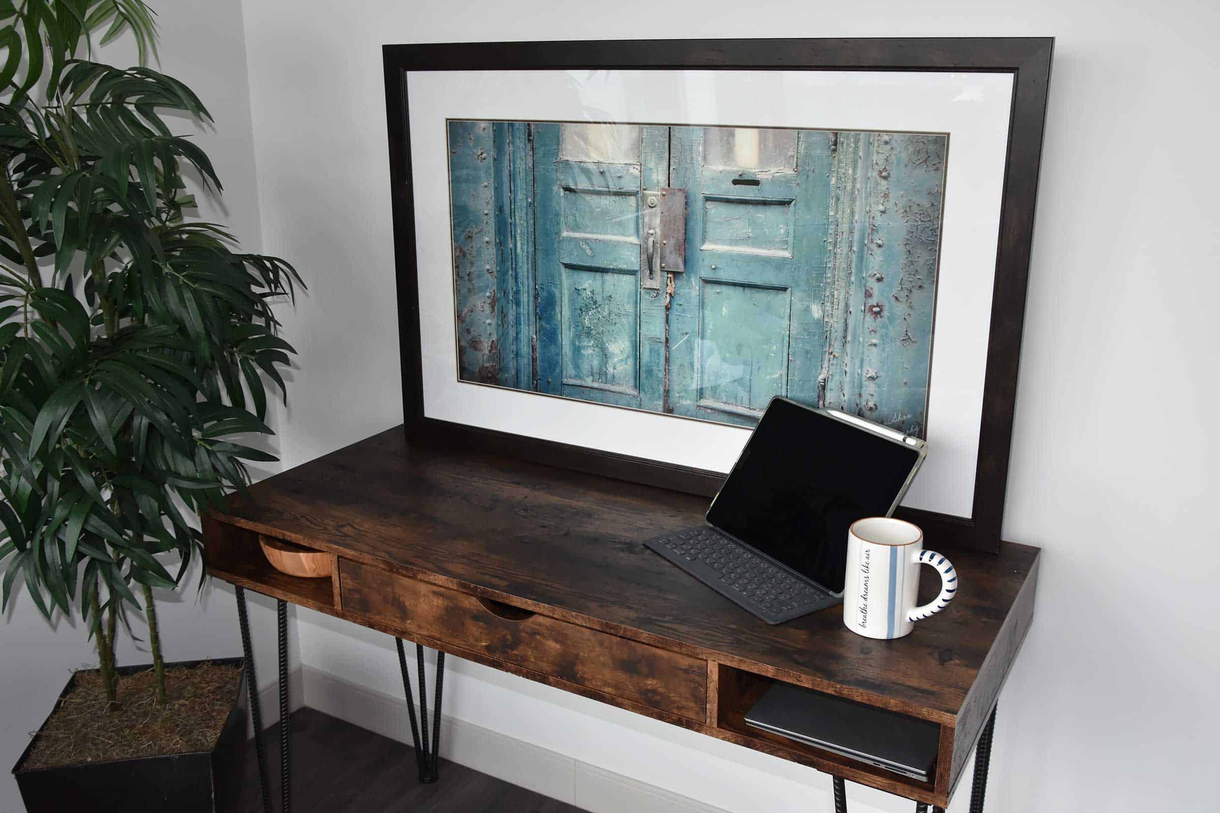 dark brown desk with laptop and mug set up as a home office. the desk sits against a wall that has a large framed photo of a blue door on it. A faux ficus tree sits in the corner next to the desk.